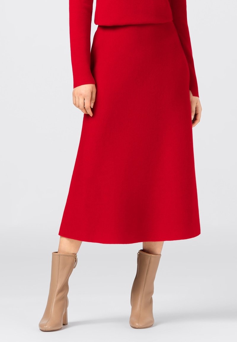 Jupe midi rouge en tissu lisse, avec une taille ajustée et une silhouette trapèze, associée à des bottes beige montant jusqu'aux genoux.