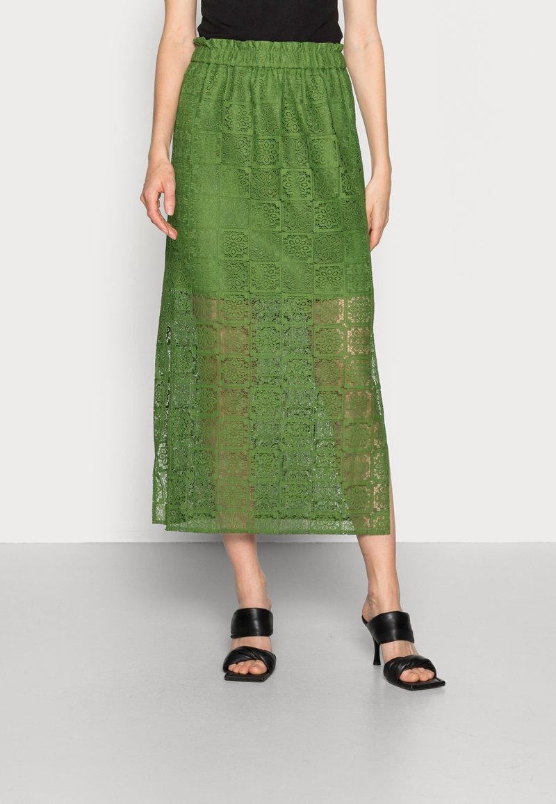 Femme portant une jupe midi en dentelle verte avec des motifs floraux carrés et des sandales noires à talons, debout devant un fond uni.