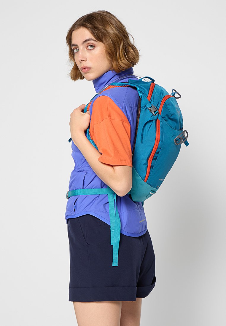 Ryggsäcken har en turkos utsida med orange accenter, en konturerad form, justerbara remmar och en ficka med dragkedja. Användaren har på sig en flerfärgad outfit.