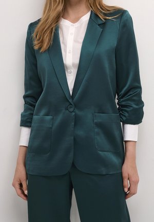 Blazer en satin teal au design ajusté, fermeture à un bouton, manches retroussées et deux poches avant, associé à une chemise blanche à boutons.