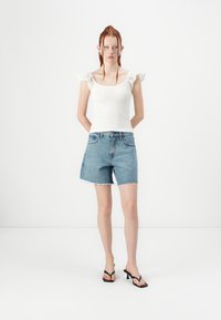 Witte, gestructureerde top met gehaakte ruffles aan de mouwen, gecombineerd met hoge, lichtblauwe denim shorts met een ruwe zoom en zwarte sandalen.