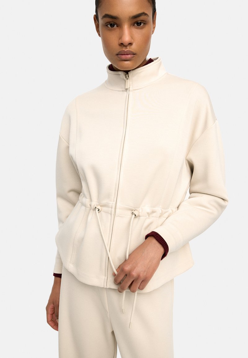 Młoda kobieta w kremowej kurtce na zamek z regulowanym w pasie sznurkiem i dopasowanych spodniach, stojąca na jasnym, jednolitym tle.