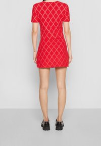 Mujer vestida con una blusa roja de mangas cortas y una falda corta a juego con un patrón de diamantes, combinada con zapatos negros de suela gruesa, de pie de espaldas.
