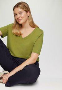Jonge vrouw met blond haar zit met gekruiste benen in een groen V-hals shirt, donkere broek en beige sneakers tegen een effen achtergrond.