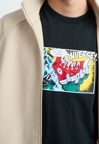 Kreslená grafika rukou držících červenou tenisku Converse s textem "Její síla je příliš velká na to, aby ji bylo možné ovládnout!" na černém tričku.