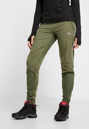 Person trägt olivgrüne Outdoor-Hose und schwarze Wanderschuhe mit pinken Akzenten, steht mit den Händen seitlich vor weißem Hintergrund.