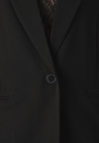 Črn blazer z gladko teksturo, ki vsebuje en sam črn gumb, klasične reverje in subtilne žepe spredaj. Pod njim je vidna čipkasta podrobnost.