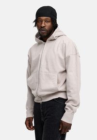 Light beige zip-up hoodie with a front pocket and ribbed cuffs, made from soft fabric. Worn with dark jeans and a black cap.