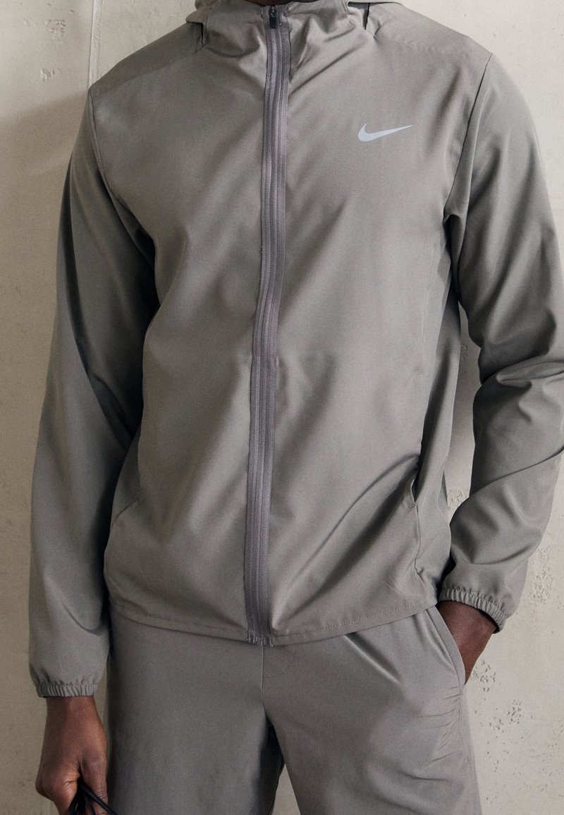 Homme portant un sweat à capuche zippé gris Nike et un pantalon assorti, debout avec une main dans la poche et tenant un objet dans l'autre main.