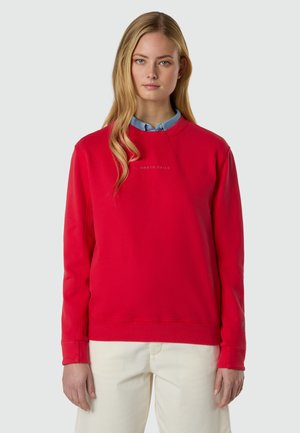 Femme blonde portant un sweat-shirt rouge vif par-dessus une chemise à col bleu et un pantalon blanc, debout devant un fond clair uni.