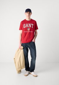 Tricou roșu din bumbac cu logo-ul "GANT", blugi albaștri, jachetă beige tip bomber ținută în mână, adidași albi cu talpă de gumă, șapcă bleumarin.
