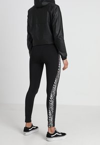 Svart kort jacka med huva, i kombination med svarta leggings med en vit mönstrad sidopanel och svarta sneakers.