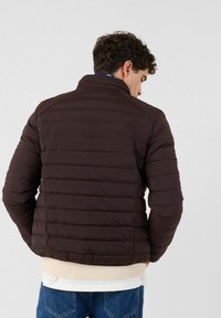 Bruine gewatteerde jas met horizontale stiksels, hoge kraag en geribbelde zoom. Draagt over een wit overhemd, gefotografeerd van achteren.