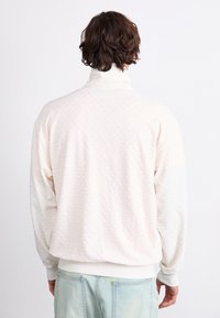 Man med medellångt brunt hår som bär en texturerad vit polotröja och ljusblå jeans, sedd bakifrån, stående mot vit bakgrund.