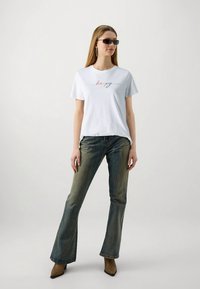 Sieviete, kas valkā saulesbrilles, baltu t-kreklu ar vārdu "laimīgs", zilas plati piegriezuma džinsus un brūnas norādītas kurpes, stāv uz neizteiksmīga fona.