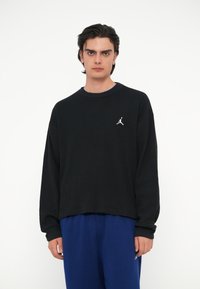 Črna pulover majica s waffle pletenjem, ki ima rebrast ovratnik in majhen bel Jumpman logotip na levem prsnem delu. Ohlapen kroj.