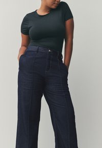 Top negru cu mâneci scurte, combinat cu pantaloni largi din denim închis, cu talie înaltă, buzunare laterale și detalii de cusături contrastante.