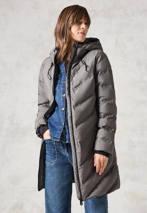Manteau gris rembourré avec capuche, doté de cordons de serrage élastiques, de poches latérales et d'une fermeture éclair. Porté par-dessus une chemise en jean et un jean bleu.