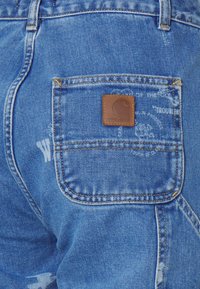 Plave traperice s kvadratnom smešnom kožnom oznakom s Carhartt logotipom, vidljivim šavovima i stražnjim džepom s blagim tekstualnim detaljem.