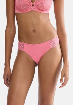 Žena v růžové krajkové podprsence a odpovídajících kalhotkách, stojící s uvolněnýma rukama podél těla na jednobarevném pozadí.