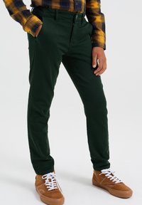 Pantaloni in cotone verde con una vestibilità slim, dotati di tasche laterali e passanti per cintura. Abbinati a sneakers in suede marrone con lacci bianchi.