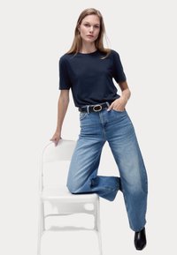 Marinblå crew-neck t-shirt matchad med ljusblå vida jeans. Svart bälte med guldkopp, modell lutad mot en vit stol. Slät tygtextur.