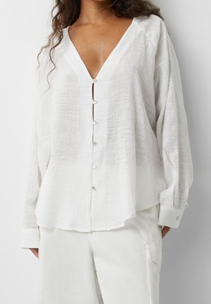 Femme portant un chemisier blanc texturé à manches longues et amples, à boutons et col en V profond, assorti à un pantalon blanc avec poche latérale.