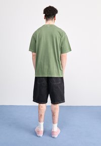 Camiseta de algodón verde con mangas cortas, combinada con pantalones cortos de mezclilla negros. Lleva puestos unos zapatos de lona rosas y calcetines blancos. Color sólido en toda la prenda.
