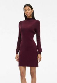 Robe en tricot côtelé bordeaux avec des manches longues, un col montant, une silhouette ajustée et une longueur au-dessus du genou.