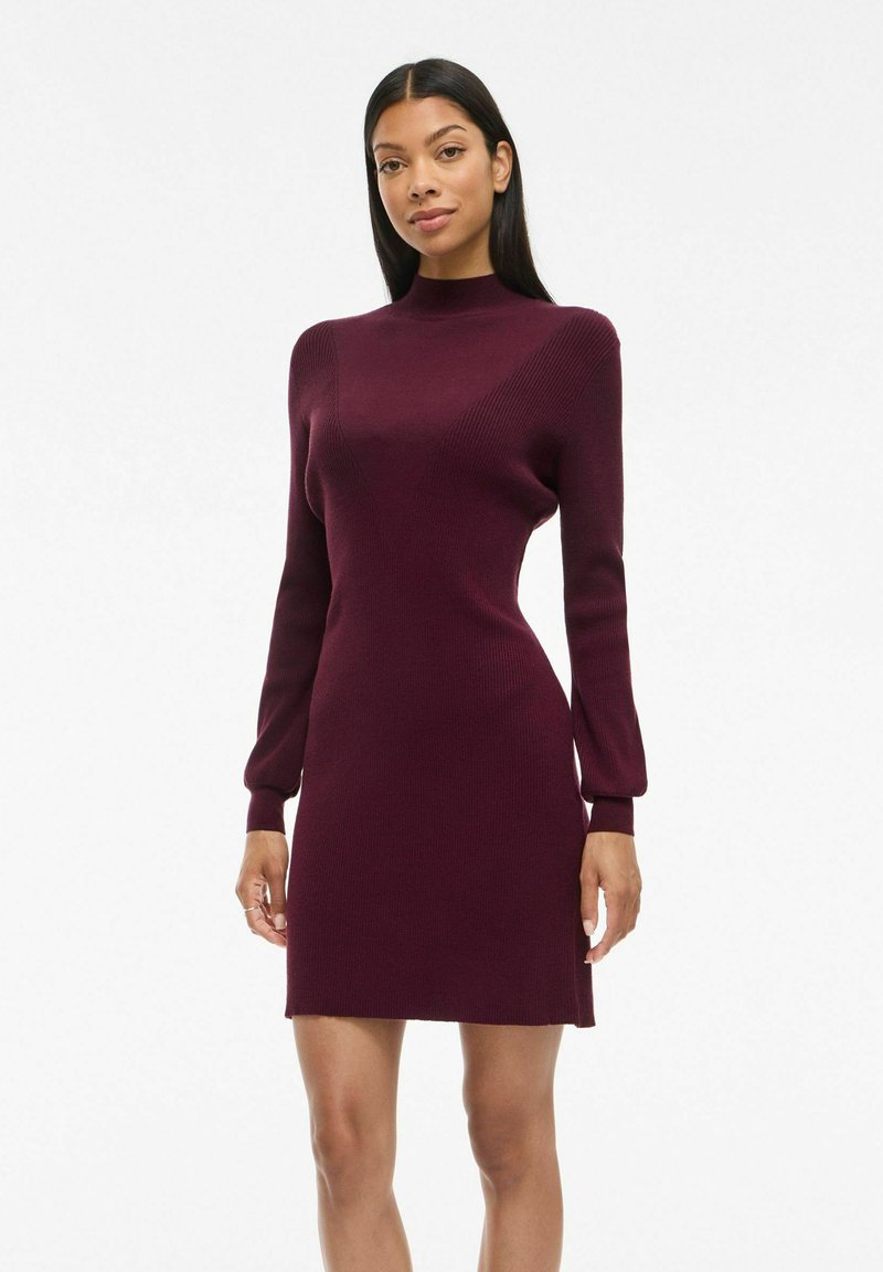 Robe en tricot côtelé bordeaux avec des manches longues, un col montant, une silhouette ajustée et une longueur au-dessus du genou.
