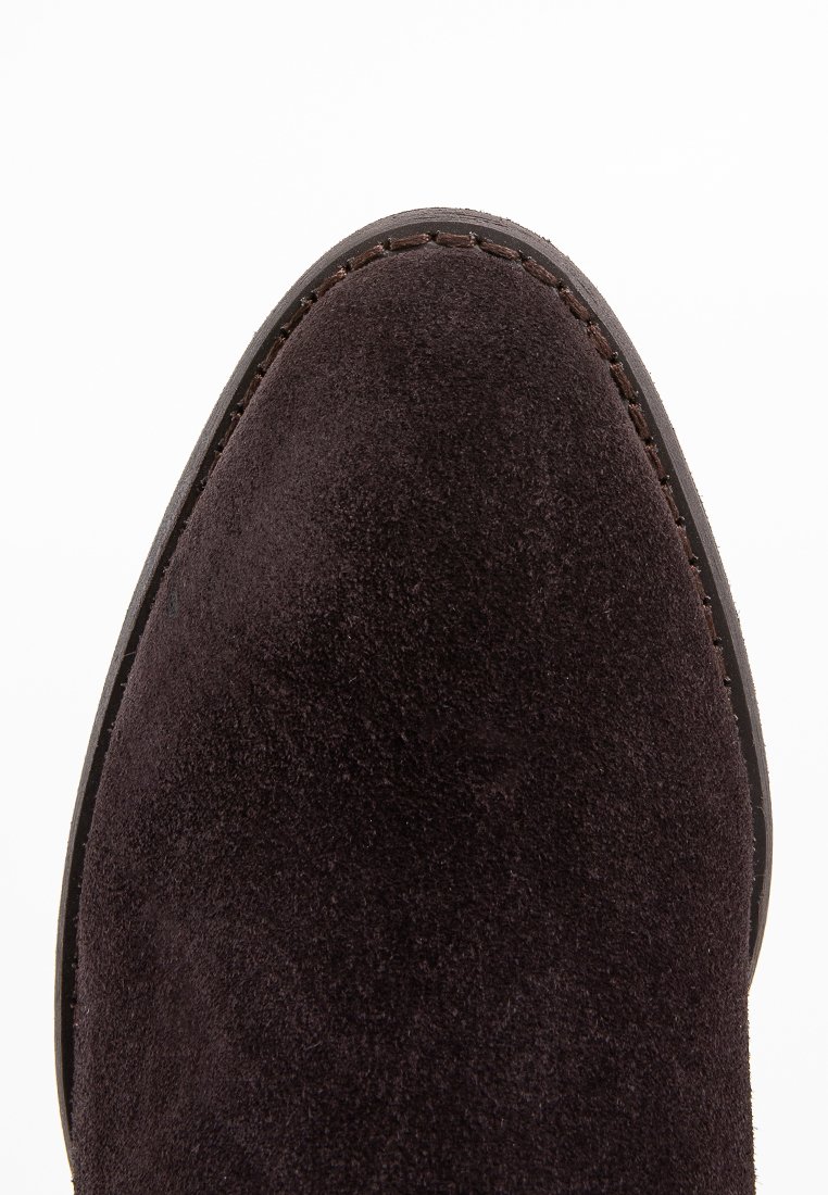 Parte superior de zapato de ante marrón con textura suave, punta redondeada y suela oscura contrastante con detalles de costura. Diseño minimalista.