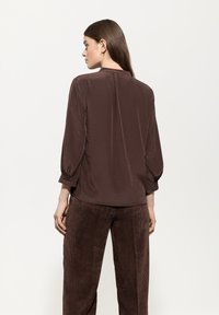 Braune, leichte Bluse mit langen, gepufften Ärmeln, glatter Textur und subtilen Glanz. Kombiniert mit beigen, weiten Cordhosen.