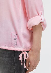 Rosa hochgekrempeltes Oberteil mit einem seitlichen Zugband, das zu einer Schleife gebunden ist, getragen mit dunkelgrauen Jeans, die einen Teil des Arms und der Hand einer Person zeigen.