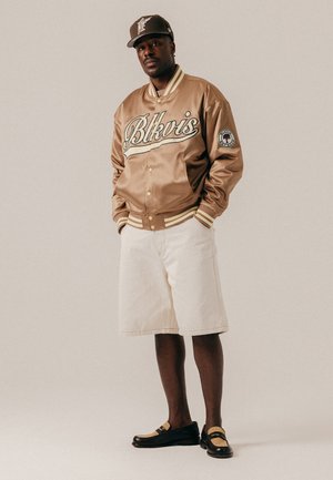 Hombre con chaqueta varsity beige con la palabra "Blkvis", pantalones cortos crema, gorra a juego y mocasines negros, de pie frente a un fondo sencillo.