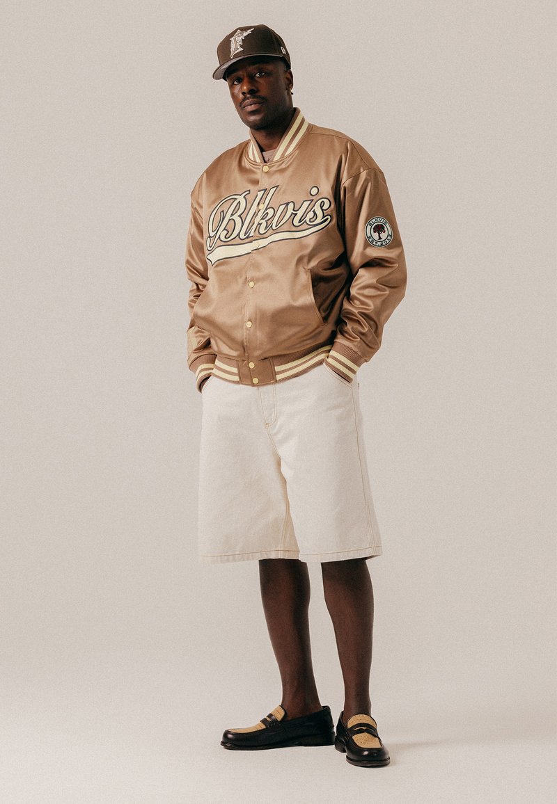 Homme portant une veste varsity beige avec l'inscription « Blkvis », un short crème, une casquette assortie et des mocassins noirs, debout devant un fond uni.