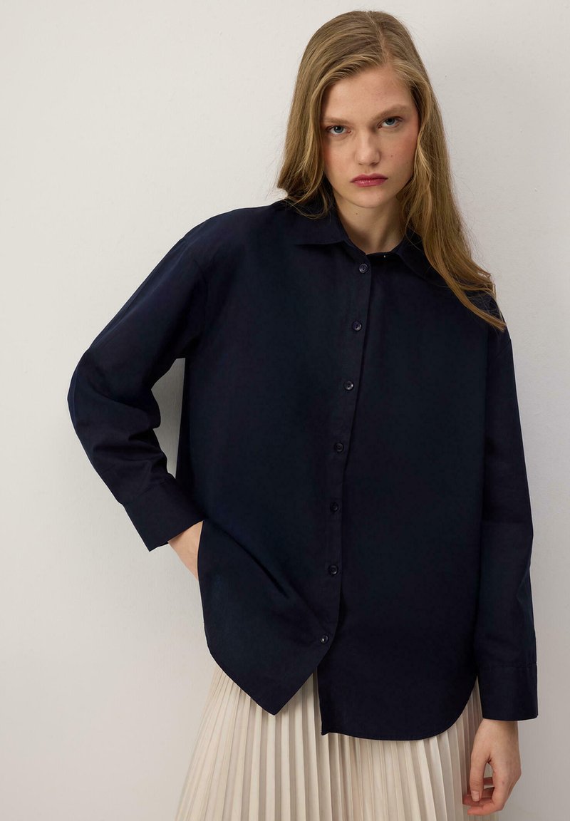 Jeune femme aux longs cheveux blonds portant une chemise ample bleu foncé boutonnée et une jupe plissée claire, debout devant un fond uni.