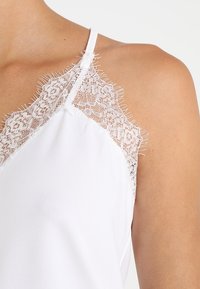 Camisola bianca con spalline sottili, caratterizzata da un rivestimento in pizzo sull'escote. Il tessuto appare liscio con dettagli frangiati lungo i bordi del pizzo.