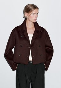 Brązowa krótka kurtka z dużymi klapami, podwójnym zapięciem, czarnymi guzikami i gładką wełnianą teksturą, noszona na jasnej bluzce i czarnych spodniach.