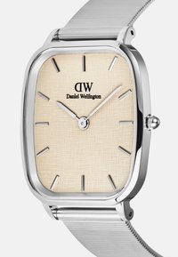 Sidabrinis stačiakampis laikrodis su tekstūruotu smėlio spalvos ciferblatu, minimalistiniais valandų žymekliais ir sidabriniu tinkleliu. Logotipas sako "Daniel Wellington."