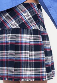 Fustă plisată în carouri în nuanțe de albastru marin, alb și roșu, cu pliuri și tiv desfăcut. Dispune de un buzunar lateral cu un mic logo al brandului. Textură moale a materialului.