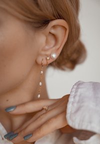 Pendientes de oro con perlas: uno con una sola perla redonda, el otro con perlas en cascada sobre una cadena. Mano con uñas azules visible en primer plano.