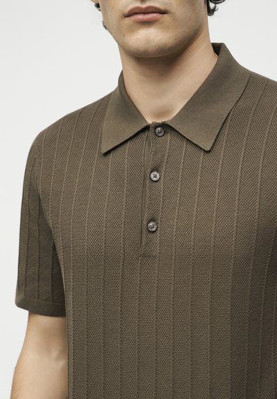 Άνδρας που φοράει καφέ πλεκτό πόλο με κοντά μανίκια, γιακά και τρία κουμπιά.