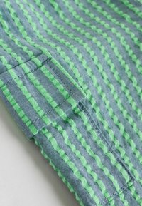 Tissu en coton avec des rayures ondulées alternant bleu et vert. La texture semble lisse avec des coutures détaillées et un bord plié.