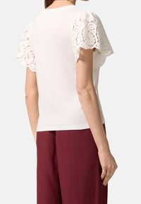 Top bianco a maniche corte con dettagli in pizzo, caratterizzato da un collo rotondo e una vestibilità morbida, abbinato a pantaloni in lino bordeaux.