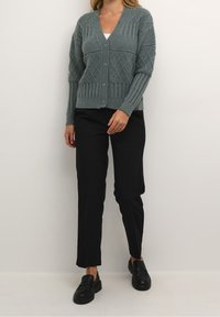 Knitted green cardigan with a V-neck and diamond patterns, featuring ribbed cuffs and hem, worn with black pants and loafers.