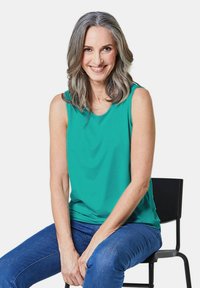 Turquoise mouwloze top in een relaxte pasvorm, gemaakt van zachte stof. Gecombineerd met blauwe jeans, op een zwarte stoel tegen een witte achtergrond.