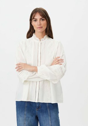 Mujer con cabello largo y castaño que lleva una blusa blanca de encaje con mangas abullonadas y vaqueros azules, de pie con los brazos cruzados contra un fondo blanco.