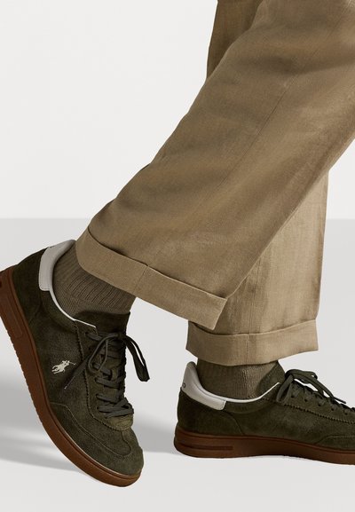 Mørkegrønne semskede sneakers med hvælvet hvid læderhælhæl, brun gummisål og sorte snørebånd, båret med tanfarvede bukser med opfoldede ben og olivengrønne sokker.