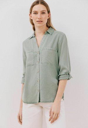 Chemise à manches longues vert clair en tissu doux, avec un col, deux poches poitrine et des manches retroussées. Texture lisse et coupe décontractée.