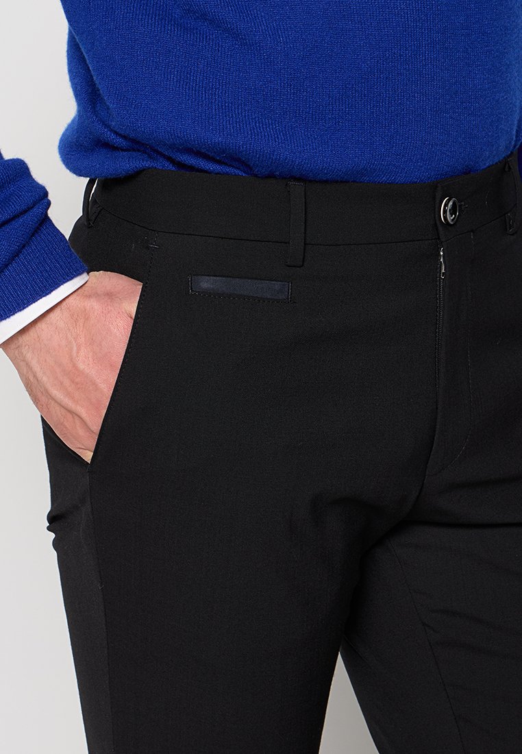Schwarze maßgeschneiderte Hosen mit slim Fit, ausgestattet mit einem versteckten Reißverschluss, Gürtelschlaufen und einer seitlichen Tasche. Das Material hat eine glatte Textur.